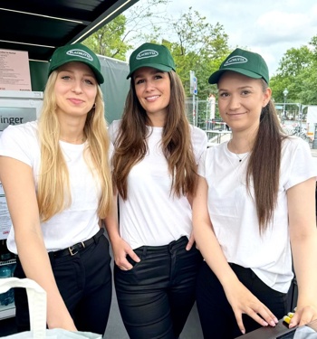 Drei Frauen in weißen Hemden und grünen Hüten stehen lächelnd an einem Veranstaltungsstand im Freien.