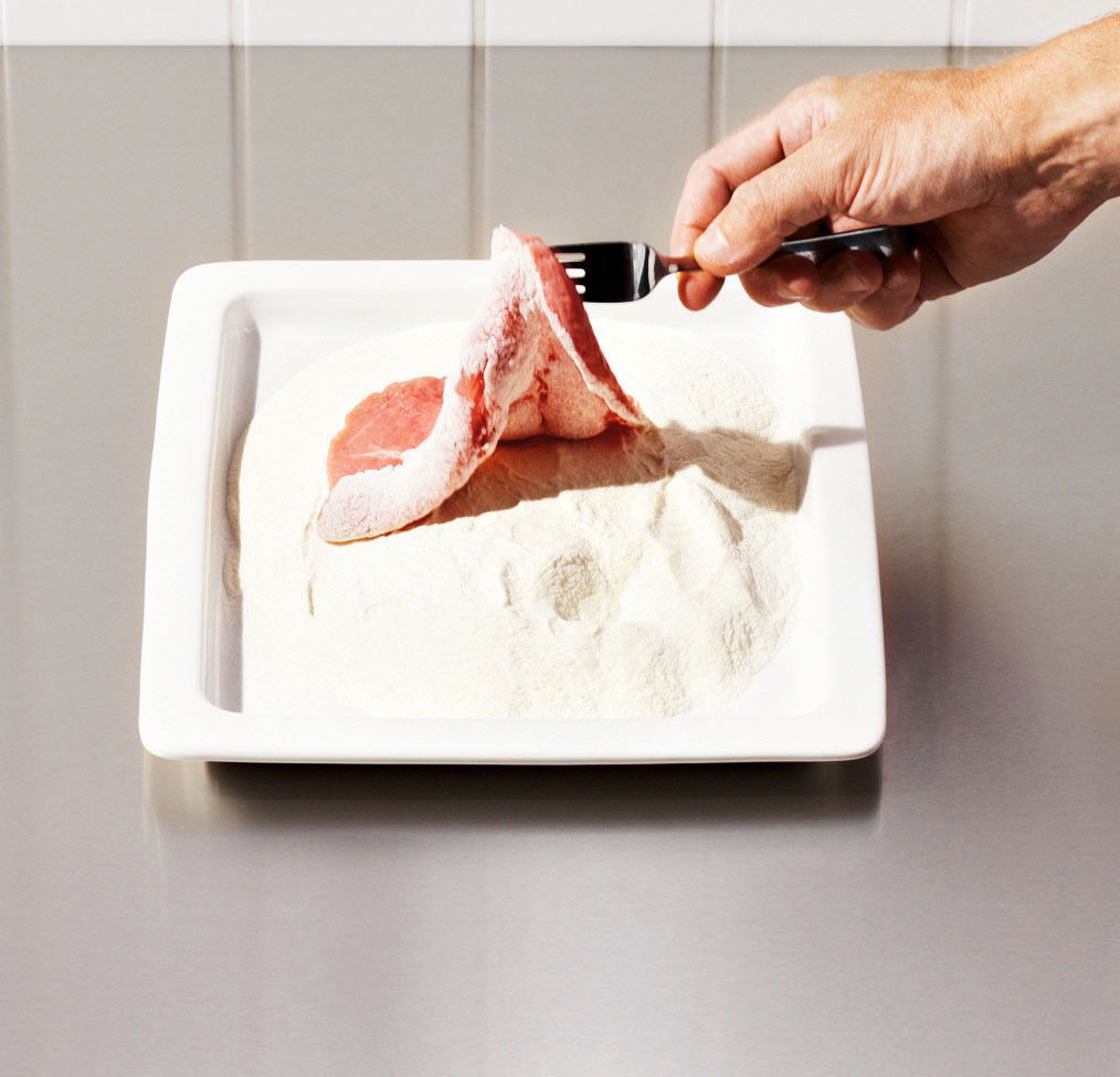 Eine Hand umhüllt mit einer Gabel ein rohes Schweinekotelett auf einem weißen Teller mit Mehl.