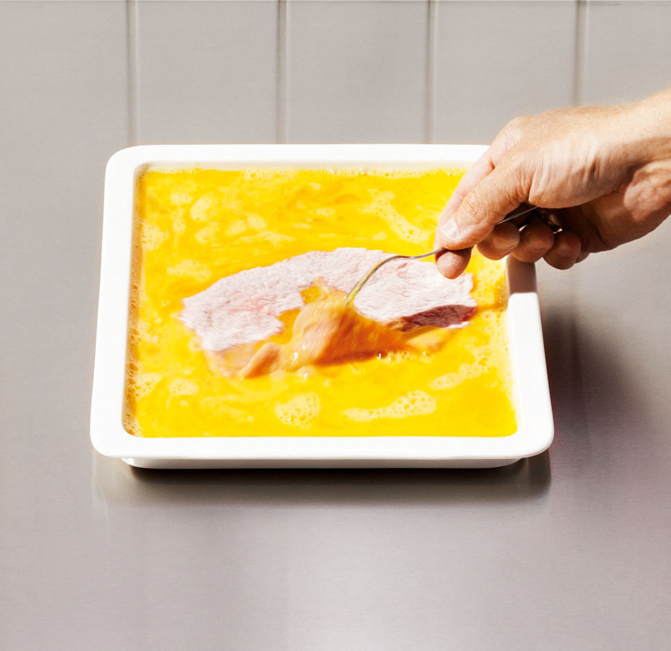 Eine Hand taucht ein Stück rohes Fleisch in eine Schale mit geschlagenen Eiern auf einer Metallfläche.