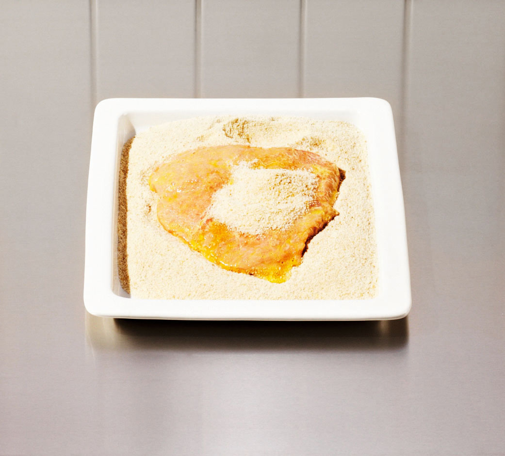 Rohes, paniertes Hähnchenschnitzel in einer weißen, mit Paniermehl gefüllten Schale auf einer Edelstahloberfläche.