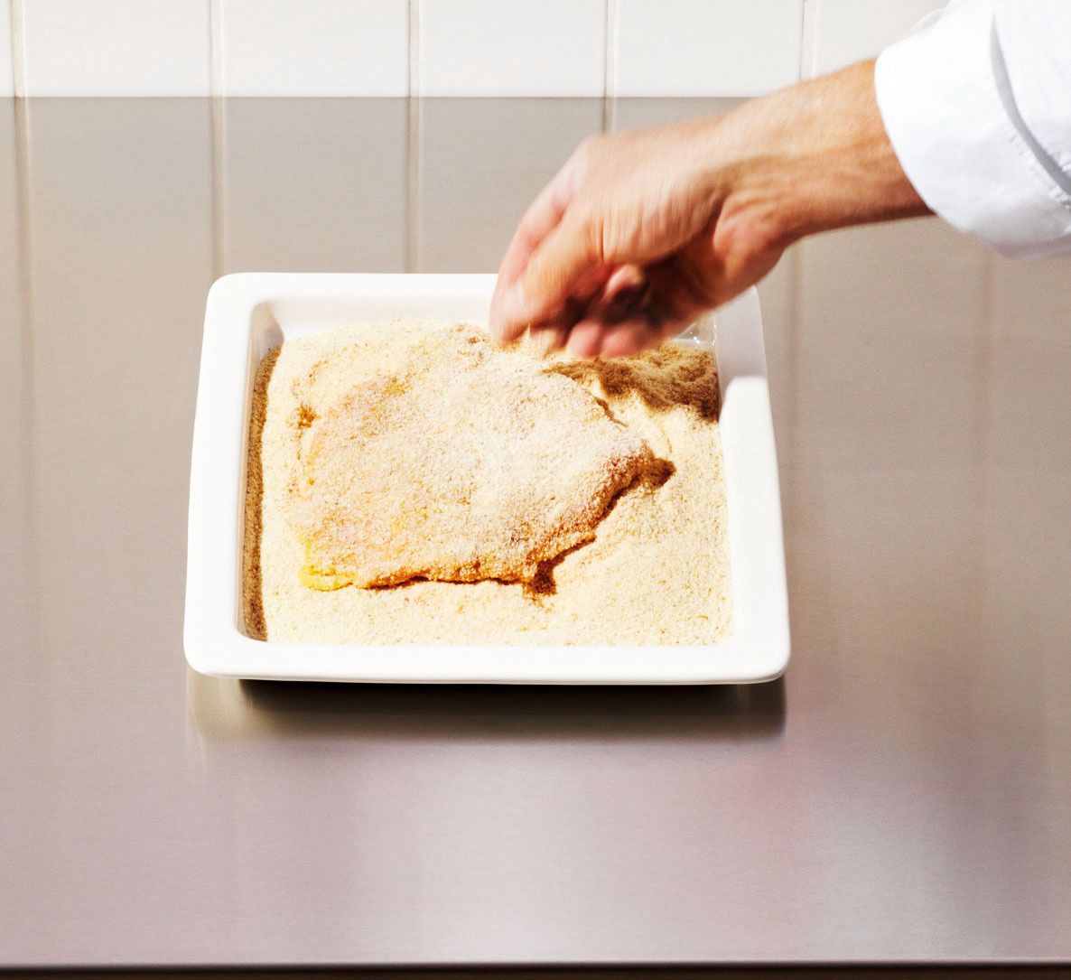 Eine Hand streut Paniermehl über ein Stück Fleisch, das in einer weißen Schale auf einem Metalltisch liegt.