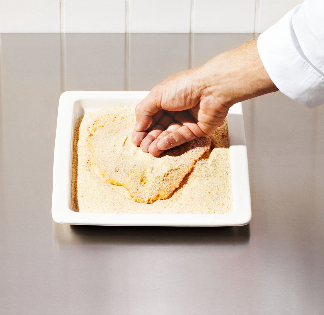 Eine Hand, die ein Stück Fleisch in einer weißen Schale auf einer Metallfläche paniert.