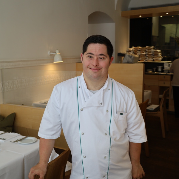 Ein Koch in weißer Uniform steht lächelnd in einem modernen, gut beleuchteten Essbereich eines Restaurants.