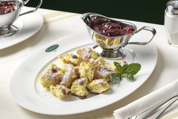 A plate of Kaiserschmarrn with powdered sugar, raisins, mint and a silver sauce boat with berry compote.