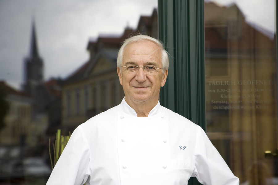 Ein älterer Mann in einem weißen Kittel steht lächelnd vor einer Glastür, in der sich Gebäude spiegeln.