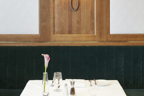 A table set for two in a restaurant with a single flower in a vase and a dark green table.