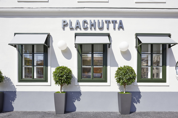 Weißes Gebäude mit zwei grün umrandeten Fenstern, grauen Markisen und Topfbäumen, Schild mit der Aufschrift PLACHUTTA.