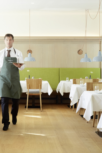Ein Kellner geht mit einem Tablett durch ein modernes Restaurant mit grünen Kabinen und Holztischen.