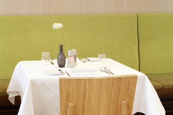 Ein Tisch für zwei Personen, gedeckt mit Geschirr, Gläsern und einer Blumenvase, vor einer grün gepolsterten Bank.