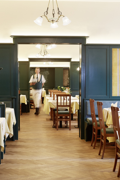 A waiter walks through a tidy restaurant with wooden chairs and tables with yellow tablecloths.