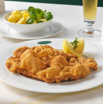Ein Teller mit paniertem Schnitzel und einer Zitronenspalte, serviert neben einer Schüssel mit Kartoffeln und Gemüse.