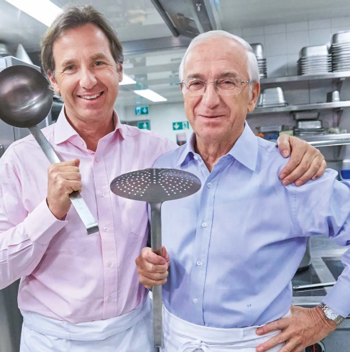 Zwei lächelnde Männer in einer Küche, die Schürzen tragen und große Küchenutensilien halten.