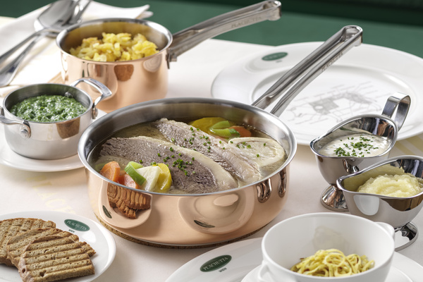 A pot of cooked beef, vegetables, sauces, noodles, greens and bread on a laid table.