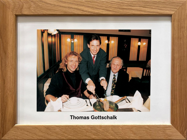 Drei Männer in formeller Kleidung sitzen an einem Restauranttisch; einer serviert das Essen. Text: Thomas Gottschalk.