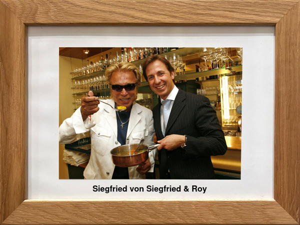 Two men in a restaurant, one with a pot and spoon, both smiling, standing in front of shelves of glasses.