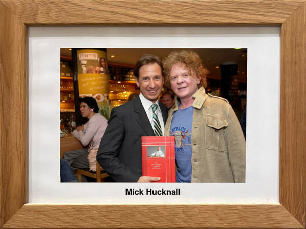 Two men pose together in a house, one holding a book in a wooden frame with the inscription Mick Hucknall.