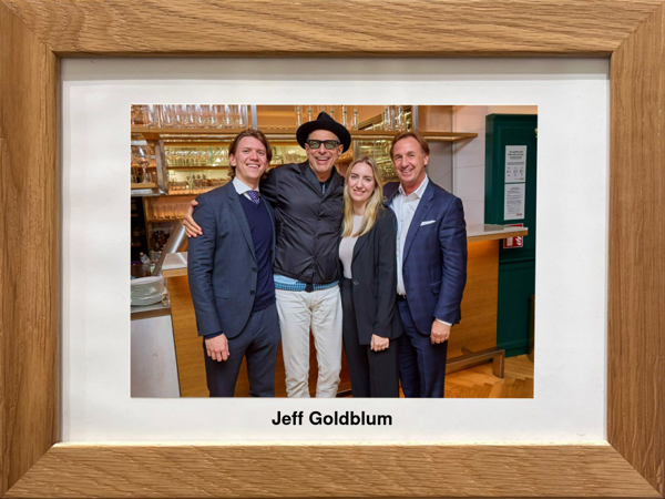 Vier Personen in Geschäftskleidung posieren mit Jeff Goldblum, der einen Hut und eine Brille trägt, in einem Restaurant.