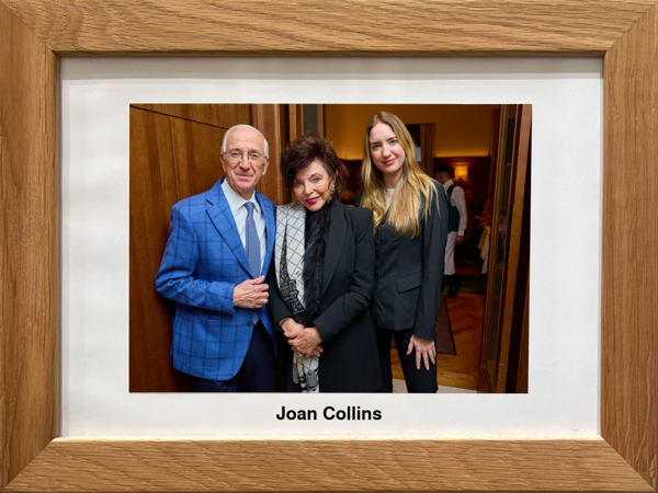 Drei Personen in formeller Kleidung posieren zusammen, unten ein gerahmtes Foto mit der Aufschrift Joan Collins.