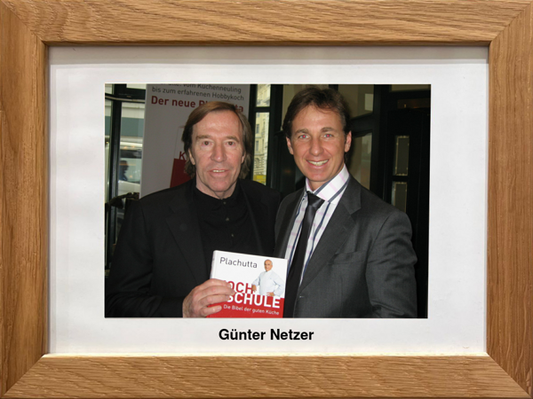 Zwei Männer in Anzügen posieren zusammen, einer hält ein Kochbuch, auf einem gerahmten Foto mit der Aufschrift Günter Netzer.
