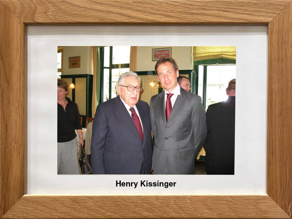 Zwei Männer in Anzügen stehen in einem Innenraum zusammen, am unteren Rand ein gerahmtes Foto mit der Aufschrift Henry Kissinger.