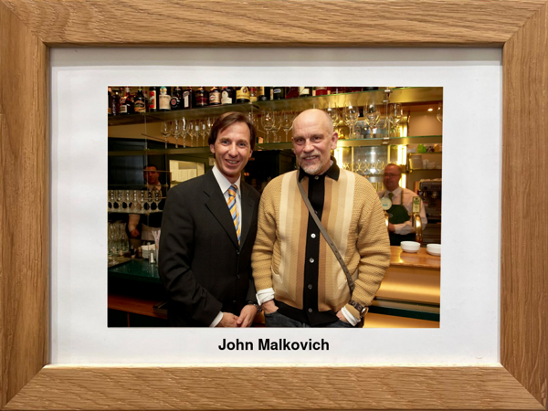 Two men pose together in a bar; one is wearing a suit, the other a beige cardigan. Text: John Malkovich.