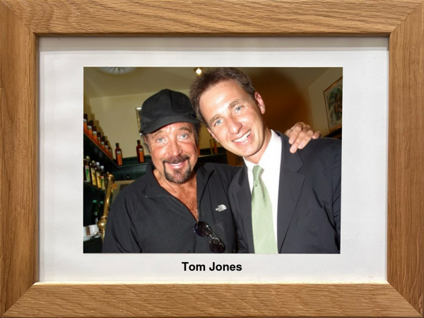 Zwei Männer lächeln sich an, der eine in schwarzer Mütze und Hemd, der andere im Anzug, gerahmtes Foto mit der Aufschrift Tom Jones.