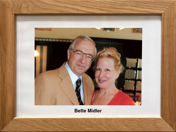 Ein lächelnder Mann in einem hellbraunen Anzug und eine Frau in einem roten Top posieren zusammen auf einem gerahmten Foto mit der Aufschrift Bette Midler.