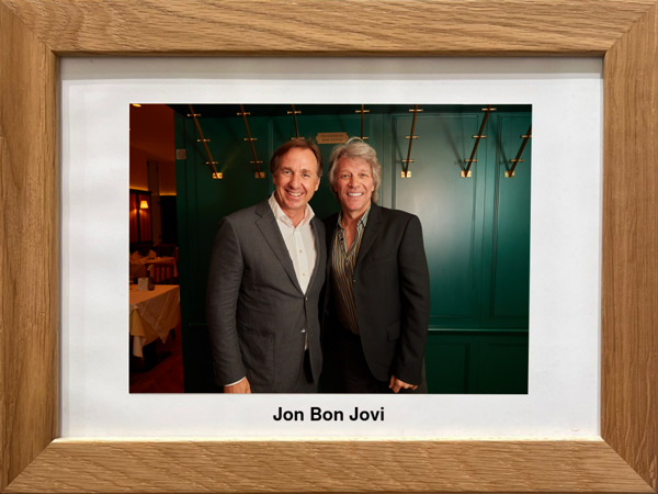 Zwei Männer in Anzügen stehen lächelnd zusammen vor einer grünen Wand mit einem gerahmten Foto mit der Aufschrift Jon Bon Jovi.