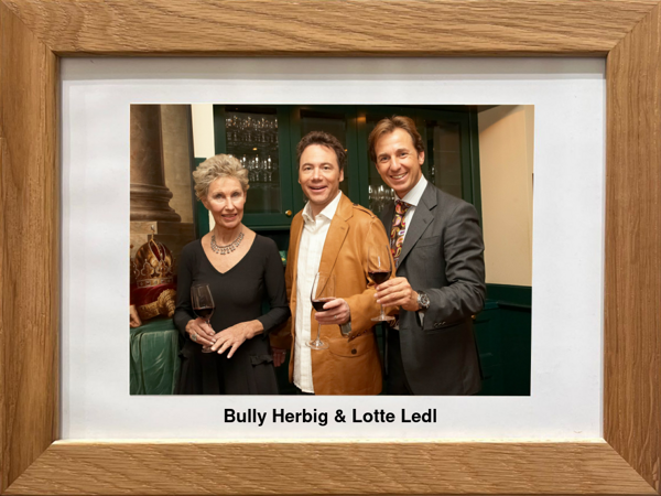 Drei förmlich gekleidete Personen stehen zusammen, halten Weingläser und lächeln in die Kamera.