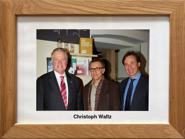 Drei Männer in Anzügen posieren zusammen in einem Innenraum; einer trägt eine Brille. Das gerahmte Bild ist mit 'Christoph Waltz' beschriftet.