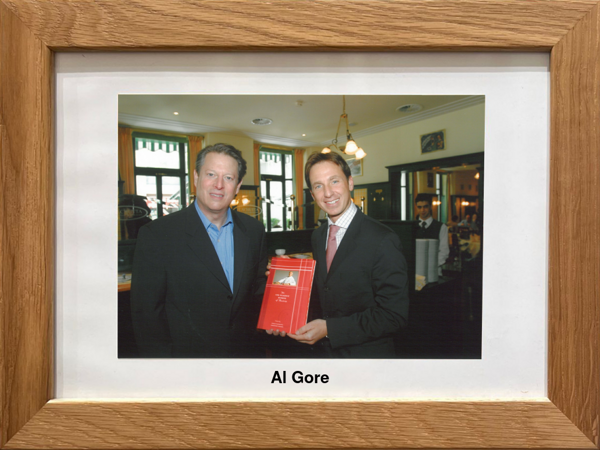 Two men in suits pose in a restaurant; one is holding a red award. Al Gore is written under the photo.