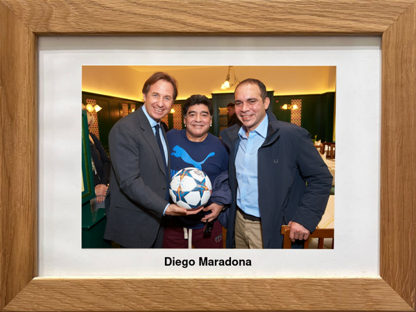 Drei lächelnde Männer in einem Haus, einer hält einen Fußball in der Hand; die mittlere Figur ist mit Diego Maradona beschriftet.