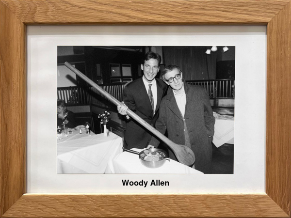 Zwei Männer posieren an einem Tisch; einer hält einen übergroßen Löffel über einen Topf. Der Text lautet Woody Allen.