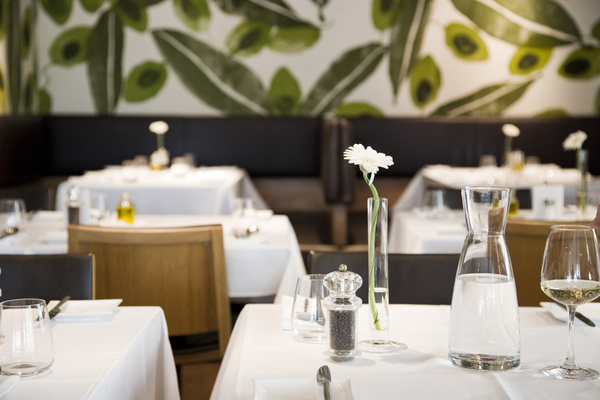 Modern restaurant with white tablecloths, simply laid tables and a green mural in the background.