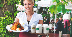 A smiling waitress holds a plate of food and a tray of beers in an outdoor restaurant.